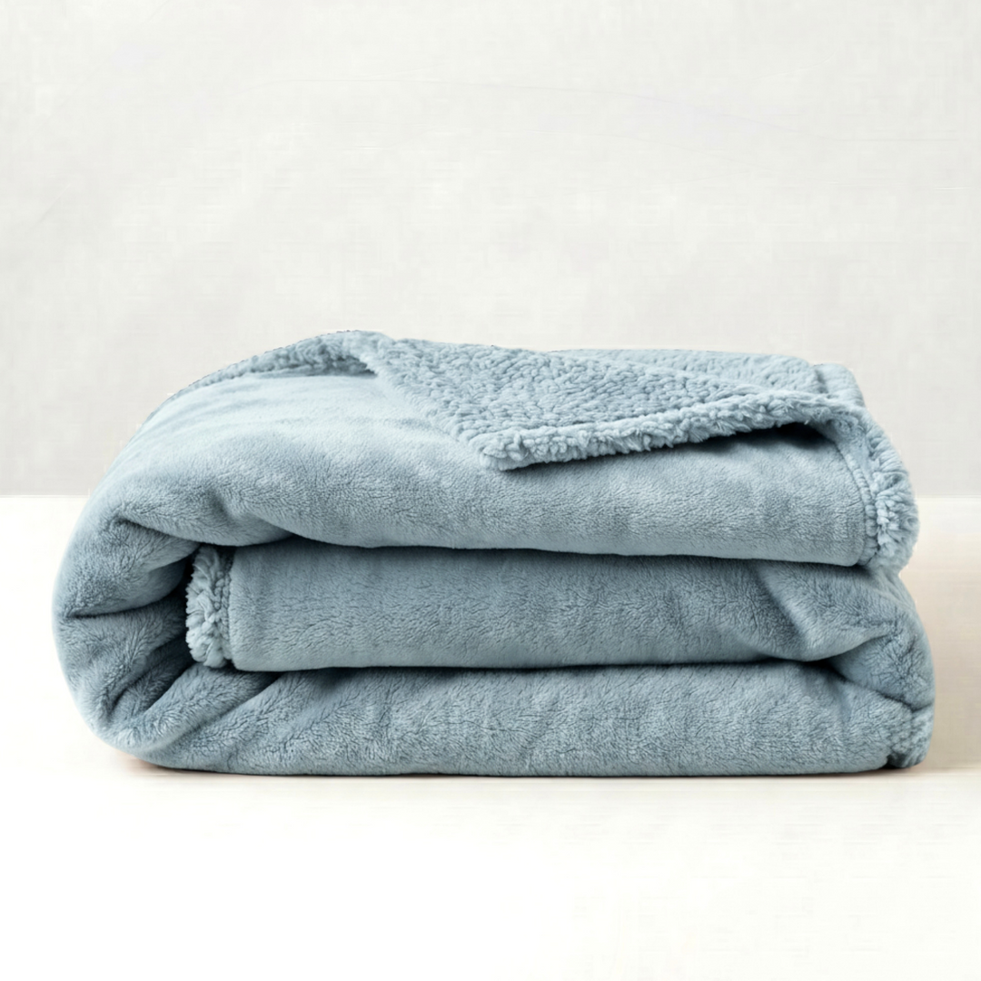 A folded dusty blue fleece blanket with a sherpa lining, displayed on a clean white surface against a light gray textured background.